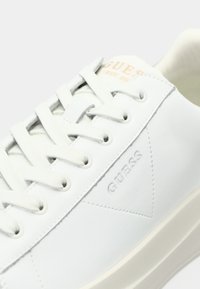 Baskets blanches Guess avec lacets blancs, logo Guess en relief sur le côté et marquage beige sur la languette.