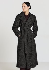 Lungo cappotto bianco e nero con motivo a fantasia, vita con cintura, ampi risvolti e tessuto strutturato. Presenta maniche lunghe e taglio dritto.