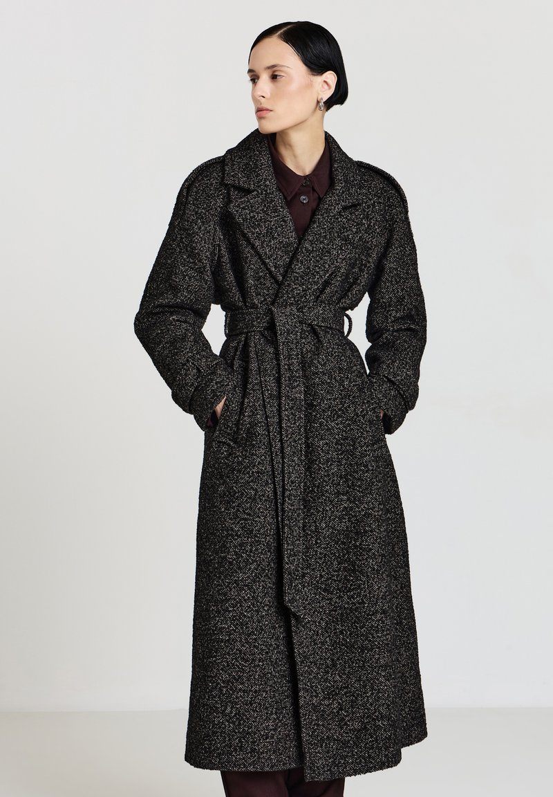 Lungo cappotto bianco e nero con motivo a fantasia, vita con cintura, ampi risvolti e tessuto strutturato. Presenta maniche lunghe e taglio dritto.