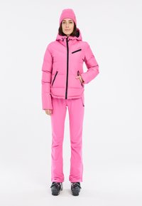 Giacca imbottita rosa con zip nere, pantaloni rosa in abbinamento e scarponi da sci neri. La giacca è dotata di cappuccio e tasche laterali.