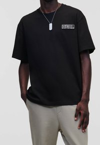 Černé tričko s volným střihem, krátkými rukávy a malým logem na hrudi. Materiál vypadá hladce a lehce.