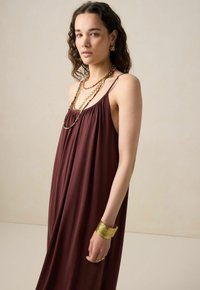 Robe sans manches bourgogne à coupe ample, confectionnée dans un tissu doux. Accentuée par des colliers superposés et un bracelet en or au poignet.