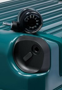 Turkooizen rollende kofferkant met een zwart wiel. Heeft een gestructureerde afwerking en een zwarte plastic behuizing met een knop en een ronde opening.