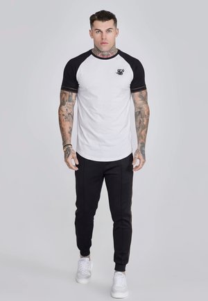 Muž s tetovaniami v bielom a čiernom raglánovom tričku, čiernych teplákoch a bielych teniskách stojaci pred jednoduchým svetlým pozadím.