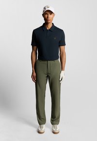 Marinblå polo med en diskret logga, olivgröna byxor och vit keps. Handskar och vita skor med bruna accenter. Släta material, avslappnad passform.