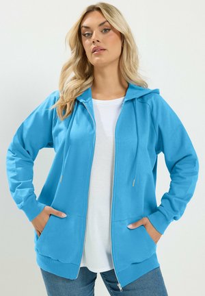 Lyshåret kvinde iført en knaldbla zip-up hættetrøje over en hvid skjorte med hænderne i lommerne, stående mod en ensfarvet lys baggrund.