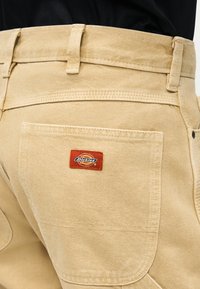 Κοντινή λήψη μπεζ παντελονιού Dickies που δείχνει την πίσω τσέπη με το κόκκινο και πορτοκαλί λογότυπο της μάρκας ραμμένο επάνω.