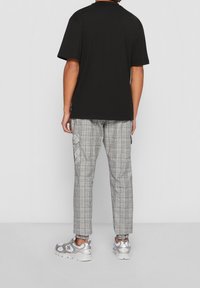 Osoba ubrana w czarną koszulę z krótkim rękawem, szare i białe spodenki w kratkę z mankietami na kostkach oraz srebrne i białe sneakersy, stojąca tyłem.