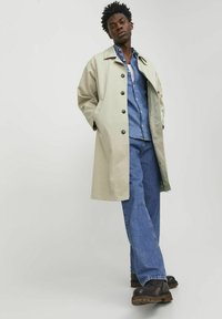 Beige regnrock med knappar, bärs över en denimskjorta. Har långa ärmar, sidofickor och avslappnad passform. Kombineras med blå jeans och mörka stövlar.