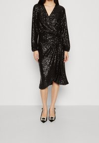 Svart sequin-inlindad klänning med lång ärm och djup V-ringning. Har en vriden front och knälång nederkant, ihop med svarta utsmyckade klackar.