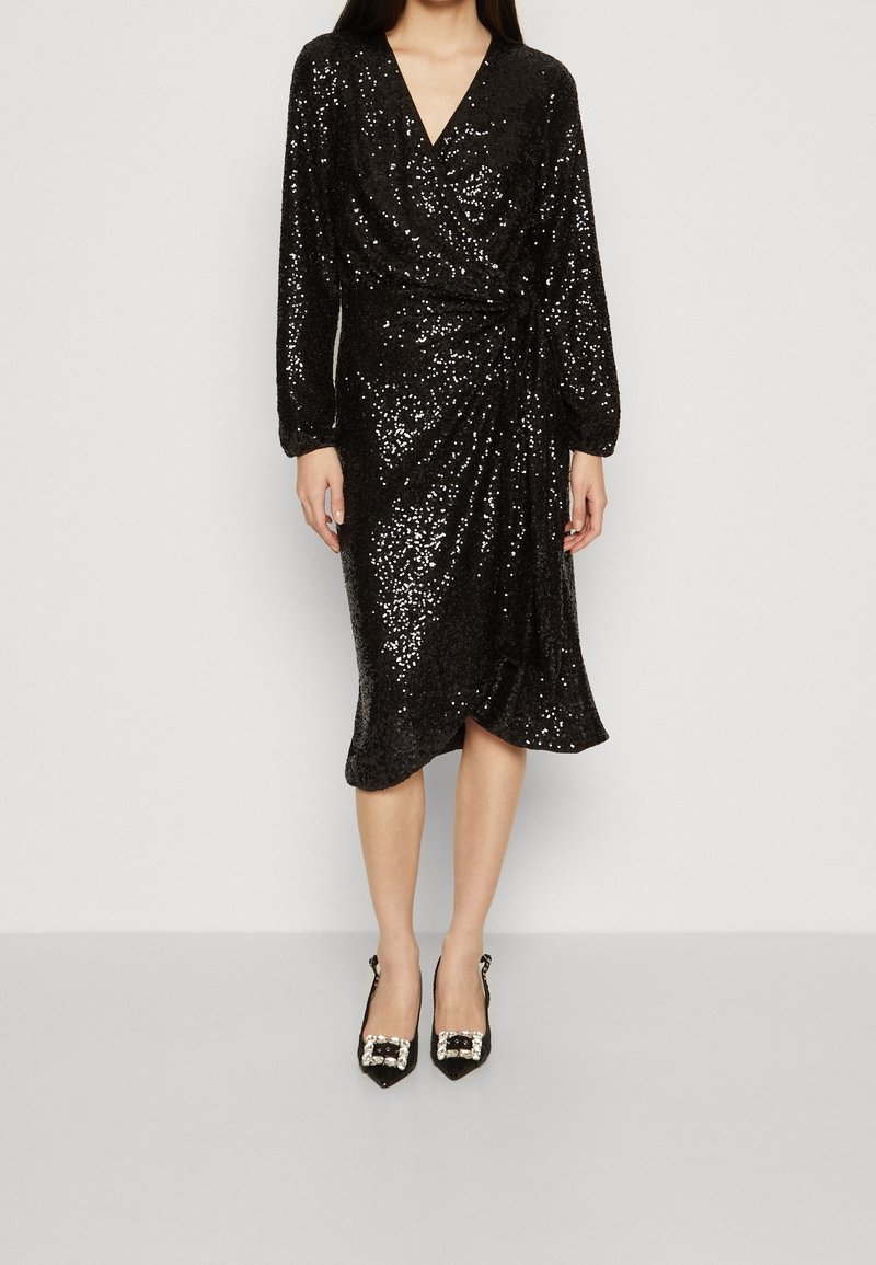 Svart sequin-inlindad klänning med lång ärm och djup V-ringning. Har en vriden front och knälång nederkant, ihop med svarta utsmyckade klackar.