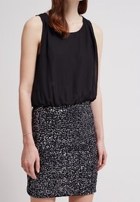 Haut noir sans manches associé à une jupe noire pailletée scintillante. Tissu lisse sur le dessus, sequins texturés en dessous, créant une silhouette ajustée.