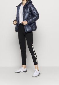 Kvinna iförd en marinblå dunjacka, vit T-shirt, svarta Champion leggings med logotyp och vita Puma sneakers med svarta mönstrade sulor.