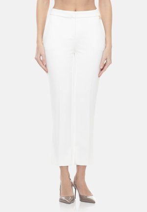 Donna che indossa pantaloni bianchi su misura alla caviglia e scarpe con tacco alto metalliche a punta, in piedi con le braccia rilassate ai lati.