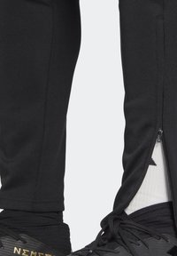 Svarta träningsbyxor med anklar som är delvis öppna, vilket avslöjar vita strumpor med en svart logotyp och svarta sportskor med guldtext.