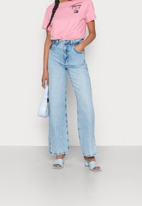 Jasnoniebieskie spodnie dżinsowe o szerokich nogawkach i wysokim stanie, w połączeniu z różową koszulką z grafiką. Dodatki to mała jasnoniebieska torebka i sandały na słupku.