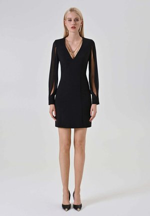 Donna bionda che indossa un vestito nero a scollo a V con maniche a fessura trasparente, collana a catena dorata e tacchi alti neri con punta, in piedi su sfondo grigio.