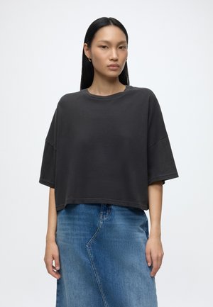 Jeune femme aux longs cheveux noirs portant un haut noir ample à manches courtes et une jupe en jean bleue sur un fond blanc uni.