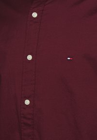 Camisa burdeos hecha de tejido suave, con un cierre de botones en la parte delantera y un pequeño logo bordado en rojo, blanco y azul sobre el bolsillo izquierdo.