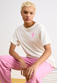 Cremefarvet kortærmet t-shirt med et pink yin-yang graphic på brystet, parret med pink sweatpants, på en siddende model mod en lys baggrund.