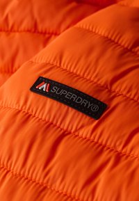 Lys orange pufferjakke i stof med quiltet tekstur. Indeholder et fremtrædende sort label med hvid og rød "SUPERDRY" branding.