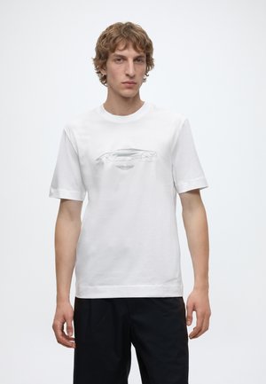 Giovane uomo con capelli castano chiaro che indossa una t-shirt bianca con uno schizzo di un'auto argentata e pantaloni neri su uno sfondo bianco uniforme.