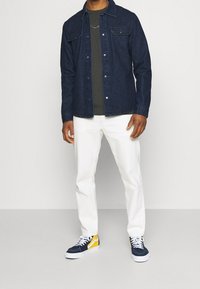 Chemise en denim foncé avec boutons-pression, t-shirt olive, pantalon blanc et baskets bleu marine/jaune avec lacets blancs. Modèle debout sur fond neutre.