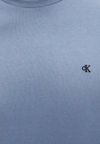 Φωτεινό γαλάζιο ύφασμα σε κοντινή λήψη με μικρό μαύρο κεντημένο λογότυπο "cK" στη δεξιά πλευρά κοντά στην περιοχή του στήθους.