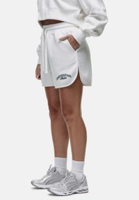Witte sweater en short set met groene tekst, voorzien van een trekkoord in de taille, zijzakken en zijsplitten. Zilveren hardloopschoenen met witte sokken.