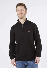 Sweat à zip noir avec un col haut, fabriqué en tissu doux. Il présente une petite patte de logo marron sur la poitrine gauche et des poignets côtelés.