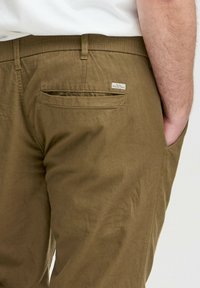 Khaki puuvillased püksid, millel on sile tekstuur. Omadusteks on tagatasku, vööloopid ja väike kangast silt tasku lähedal.