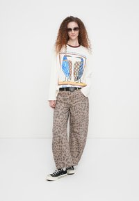 Μακρυμάνικο κρεμ πουκάμισο με σχέδιο πουλιού, συνδυασμένο με παντελόνι φαρδύ με λεοπάρ σχέδιο. Φορεμένο με μαύρα αθλητικά παπούτσια και γυαλιά ηλίου.