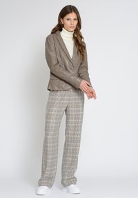 Taupe läderblazer med en knapp, kombinerad med benvita och svarta rutiga vida byxor. Modellen bär en krämfärgad polotröja och vita sneakers.