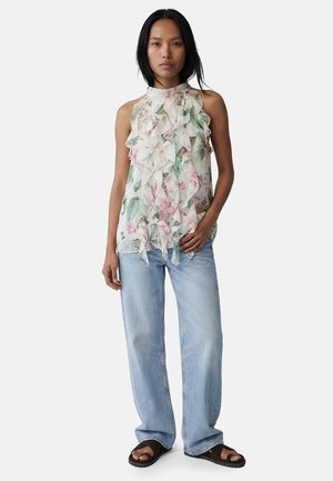 Vrouw die een mouwloze gebloemde gerimpelde blouse met hoge hals draagt, lichtblauwe losse spijkerbroek en donkere sandalen, staand met hand in de zak.