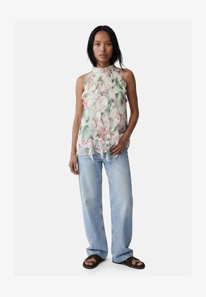 Mujer con blusa sin mangas con volantes florales y cuello alto, vaqueros sueltos azul claro y sandalias oscuras, de pie con la mano en el bolsillo.