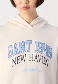 Person wearing a light beige hoodie with "GANT 1949 NEW HAVEN" in blue and black text on the chest, close-up on upper torso and lower face.
