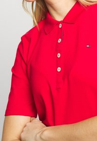 Polo rouge en tissu respirant. Il présente un col classique, des manches courtes et une patte de boutonnage avec des boutons de couleur claire.