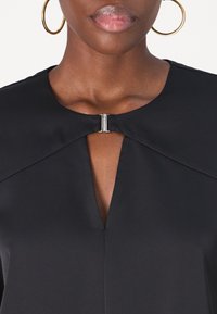 Blouse noire à col montant, avec une découpe en forme de goutte et un accent métallique au niveau du col. Tissu lisse et léger.