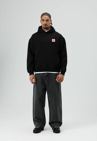 Zwarte hoodie met capuchon en voorzak, voorzien van een roze logopatch, gecombineerd met losse grijze spijkerbroek en zwarte schoenen met roze accenten.