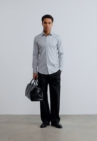 Άνδρας που στέκεται μπροστά σε απλή τοίχο, φοράει γκρι πουκάμισο με κουμπιά, μαύρο παντελόνι με φαρδιά μπατζάκια, μαύρα παπούτσια και κρατάει μια μαύρη δερμάτινη τσάντα με υφή.