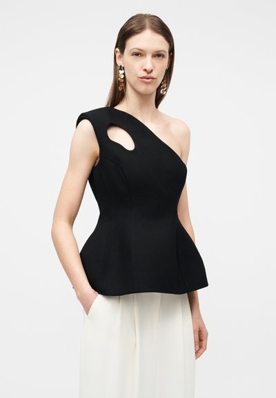 Femme portant un haut noir à une épaule avec une découpe près de l'épaule, associé à un pantalon blanc à jambes larges et des boucles d'oreilles dorées.