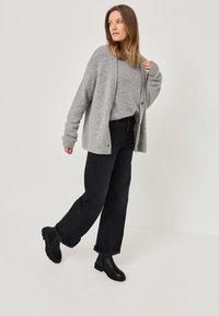 Šedý pletený kardigan s uvolněným střihem, nošený přes odpovídající šedý svetr, v kombinaci s černými širokými džínami a černými kotníkovými botami.