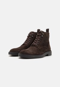 Bocanci scurți din suede maro, cu o textură netedă, vârf rotund, sistem de șireturi și o talpă din cauciuc negru. Prezintă o bucată de prindere pe spate.