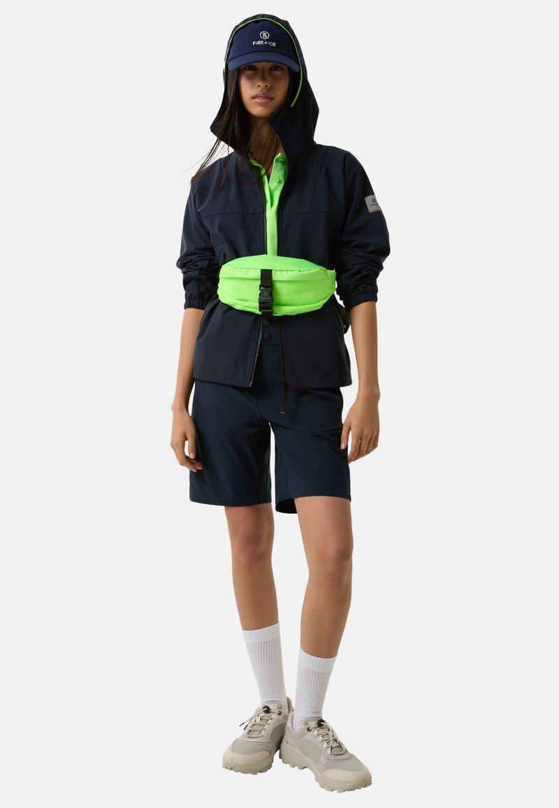 Marineblaue Kapuzenjacke mit neon grünen Akzenten, kombiniert mit marineblauen Shorts, einer neonfarbenen Gürteltasche und beigefarbenen Sneakers mit strukturierten Sohlen.