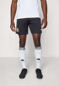 Svarte sportsshorts med gulllogo på siden, kombinert med knelange hvite sokker med svarte striper og svarte joggesko.