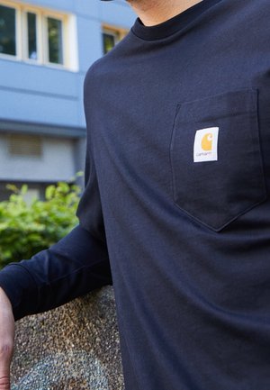 Homme portant une chemise noire à manches longues avec un logo Carhartt sur la poche poitrine, appuyé sur une surface en pierre à l'extérieur.
