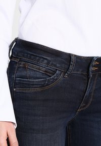 Jeans de mezclilla oscura con un diseño ajustado, ligero desgaste, costuras naranjas y detalles de bolsillos clásicos. Llevados con una camisa blanca.