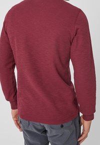 Person, der einen langärmligen, burgunderfarbenen strukturierten Pullover und graue gestreifte Hosen mit Rückentaschen trägt, von hinten betrachtet.