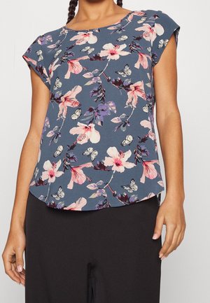 Kobieta ubrana w niebieską bluzkę z krótkim rękawem z różowo-fioletowym kwiatowym i motylim nadrukiem, w połączeniu z czarnymi spodniami.