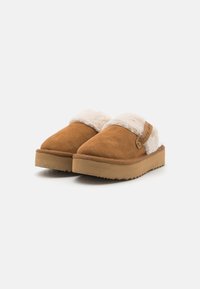 Hnědé semišové pantofle s fluffy béžovým podšíváním a texturovanou béžovou podrážkou. Obsahují boční štítek s logem na svrchní části. Design se zaoblenou špičkou.